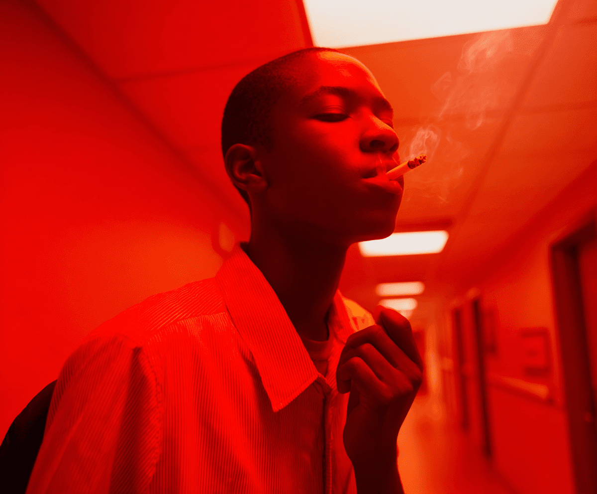Young man in a corridor washed in red emergency light, exhaling smoke, pale shirt, eyes closed