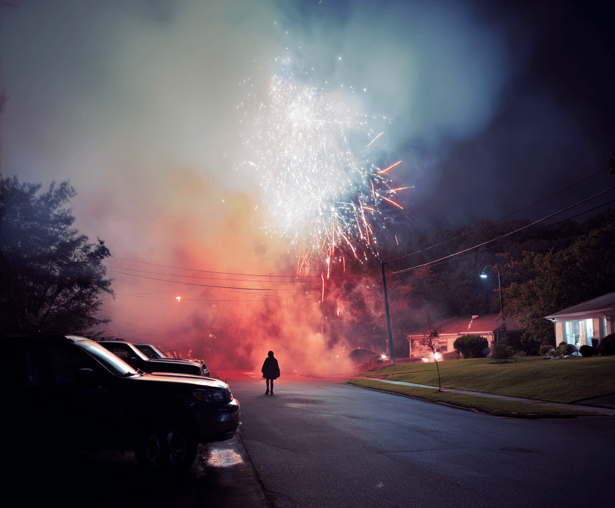 Suburban American street at night with fireworks erupting low in the sky behind houses, a lone silhouette watching
