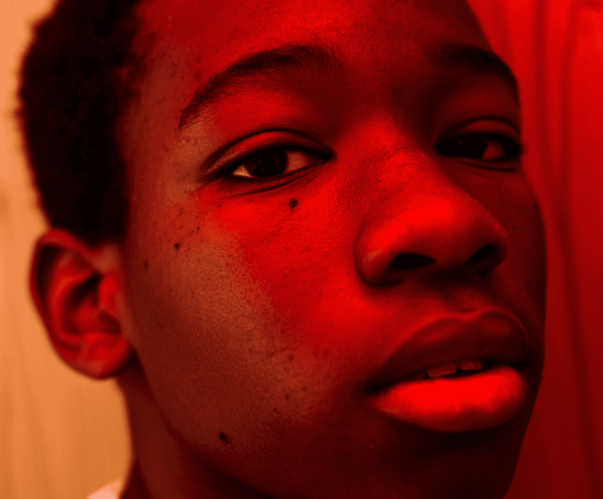Tight close-up of a young man's face under deep red light, eyes lifted, soft skin texture
