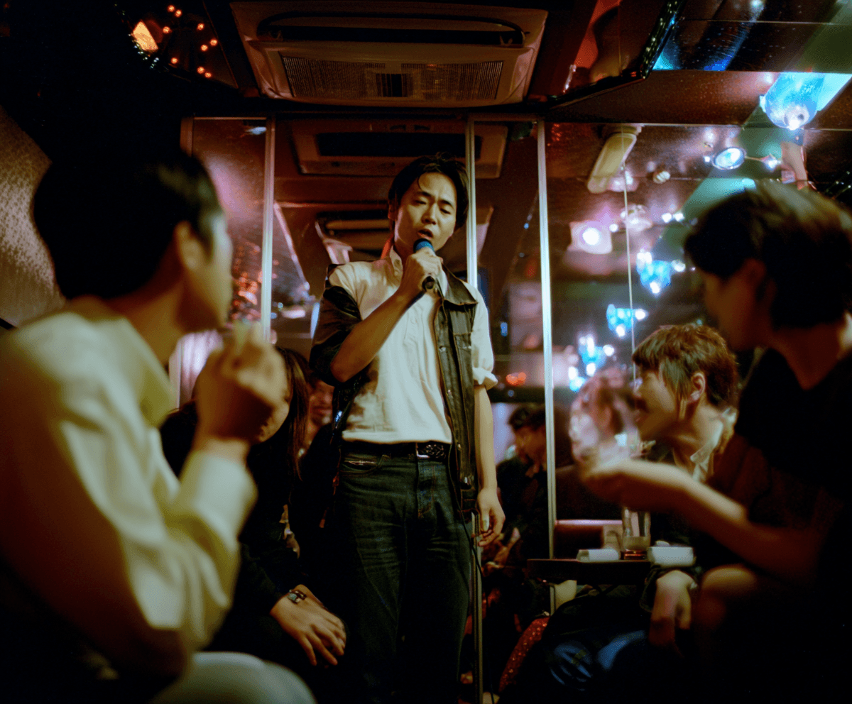 Inside a Korean karaoke party room, a young man standing center with a microphone, friends seated under string lights