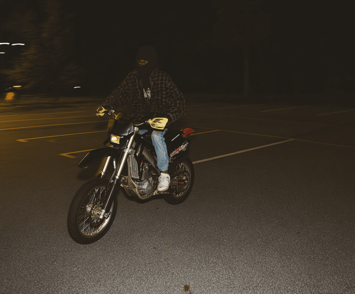 A masked motorcyclist on a dirt bike on a wet dark suburban road at night, single headlight